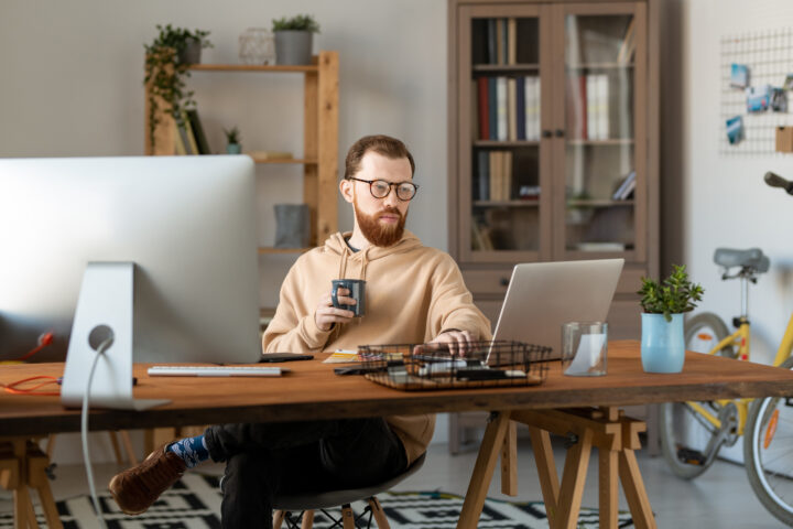 Konzentrierter freiberuflicher Designer mit Brille sitzt am Holztisch im Homeoffice und nutzt Computer beim Kaffeetrinken