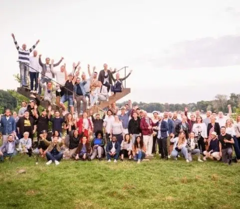 Eine Große Gruppe Von Menschen Posiert Gemeinsam Im Freien Auf Grünem Gras, Einige Stehend, Andere Sitzend Auf Oder Um Eine Wendeltreppe Herum, Alle Lächeln Und Heben Die Arme, Mit Bäumen Und Himmel Im Hintergrund.