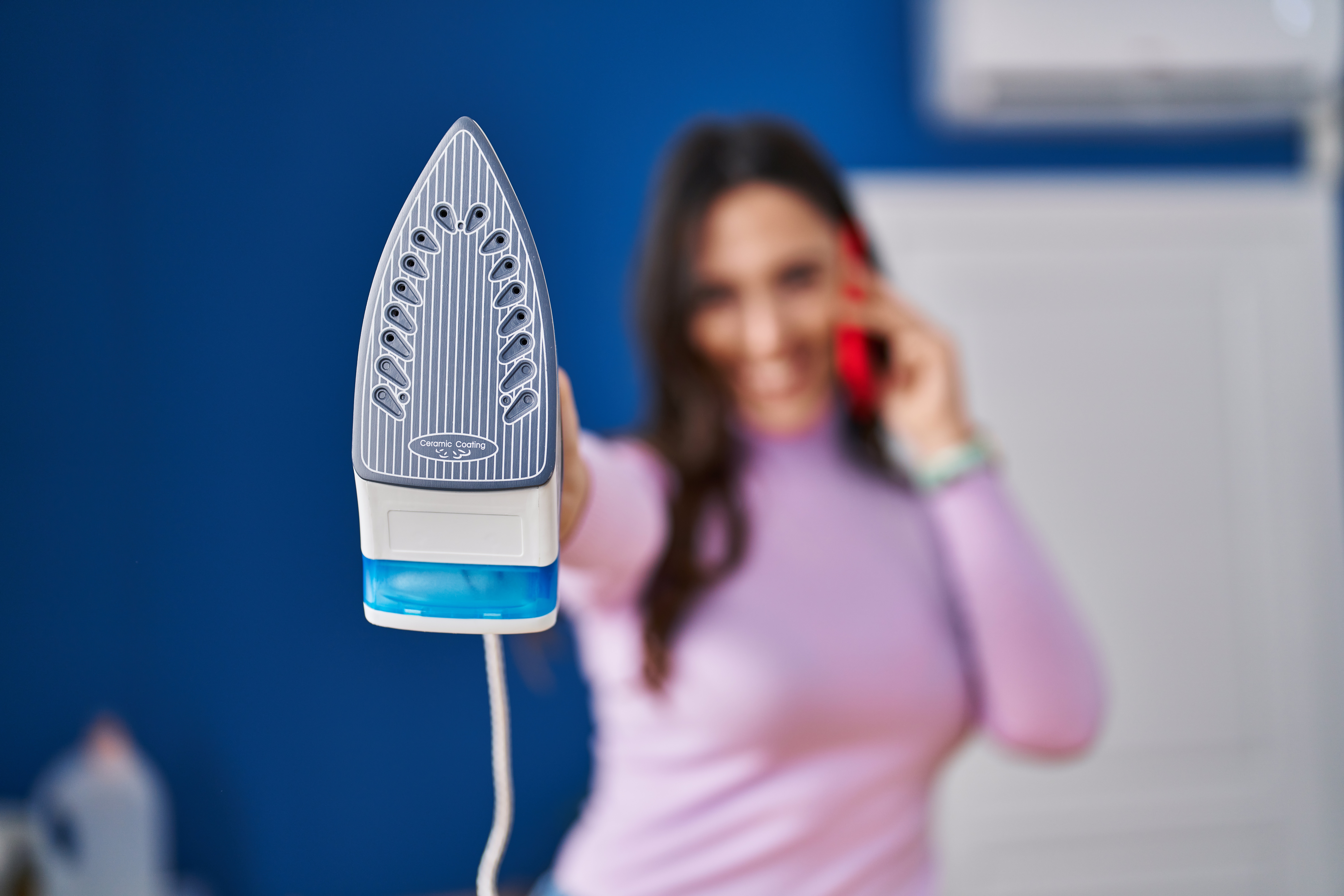 Bügeleisen im Vordergrund, Frau telefoniert im Hintergrund. Thema: Reinigung, putzen.