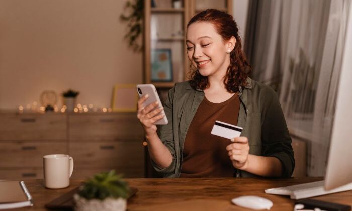 Frau am Schreibtisch mit Smartphone und Kreditkarte, lächelnd. Thema: Online-Shopping.