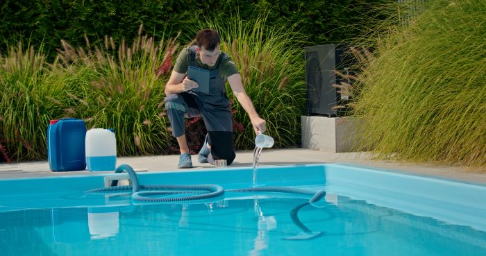 Pool Technician Pouring Liquid Into Pool While Using Tablet With Chemical Containers