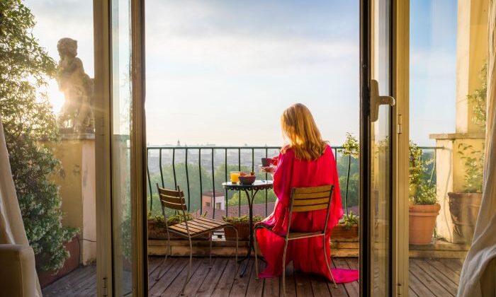 Frau in rotem Kleid frühstückt auf Balkon mit Aussicht, umgeben von Pflanzen.