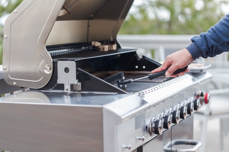A Person In A Blue Sleeve Cleans An Open Stainless Steel Grill With A Brush, Highlighting Grill Knobs. The Scene Suggests Outdoor Preparation.
