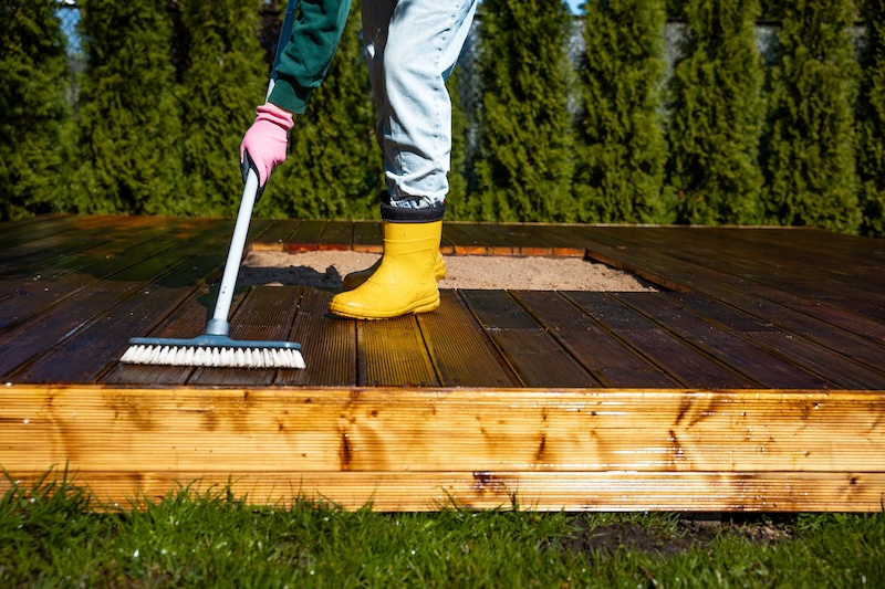Person In Yellow Boots And Pink Gloves Scrubs A Wooden Deck With A Brush. Green Trees In The Background, Sunny And Outdoors. Home Maintenance Scene.