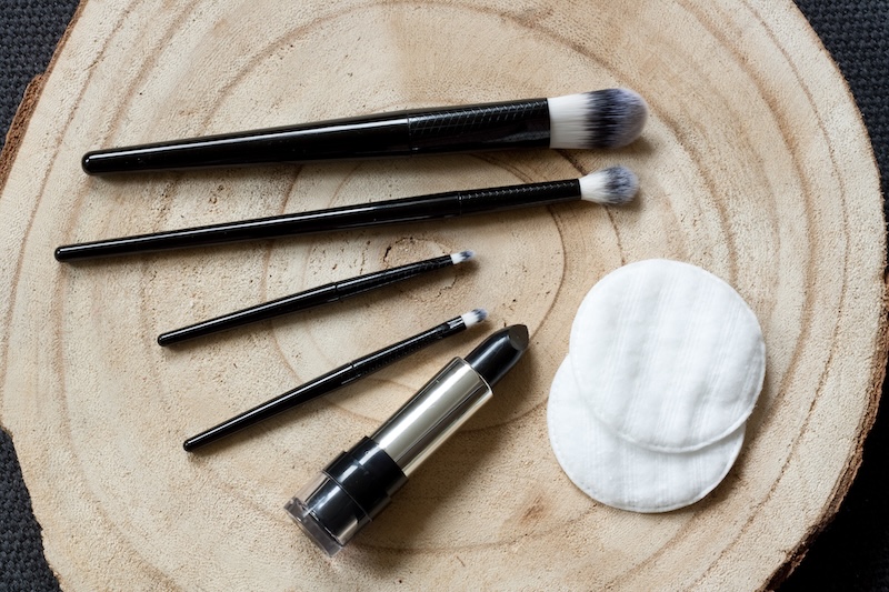 Ein Satz MakeUpPinsel Und Ein Schwarzer Lippenstift Sind Neben Weißen Wattepads Auf Einer Holzoberfläche Angeordnet. Die Szene Vermittelt Ein Gefühl Von Eleganz Und Schönheit.