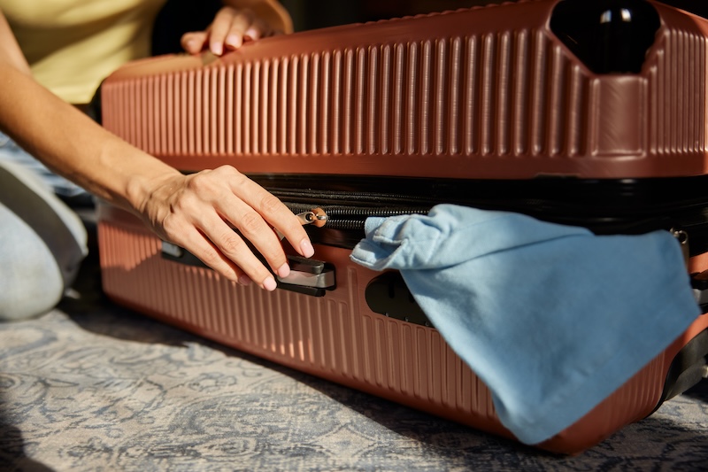 A CloseUp Of Hands Zipping A Brown Suitcase With A Blue Shirt Sticking Out. Warm Lighting Suggests A Travel Preparation Scene.