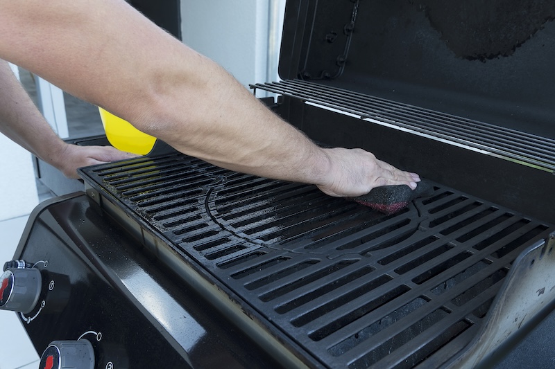 A Person Cleans A Black Grill Grate With A Scrubber Outside, Showing Attention To Detail. The Scene Conveys Cleanliness And Preparation.