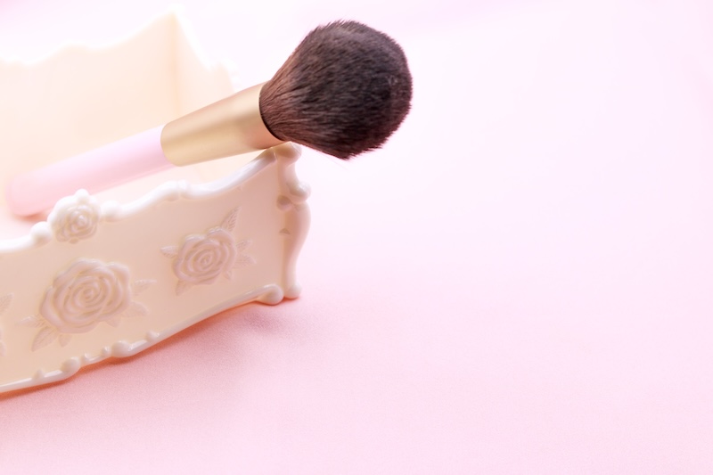 Ein MakeUpPinsel Mit Rosa Griff Ruht Auf Einem Verzierten, Cremefarbenen Halter Mit Rosenmotiven Vor Einem Zartrosa Hintergrund Und Vermittelt Einen Zarten, Femininen Ton.