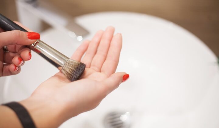 Eine Person reinigt einen Make-up-Pinsel, indem sie ihn über einem Waschbecken auf ihrer Handfläche reibt. Sie haben rot lackierte Nägel und die Atmosphäre ist ordentlich und konzentriert.