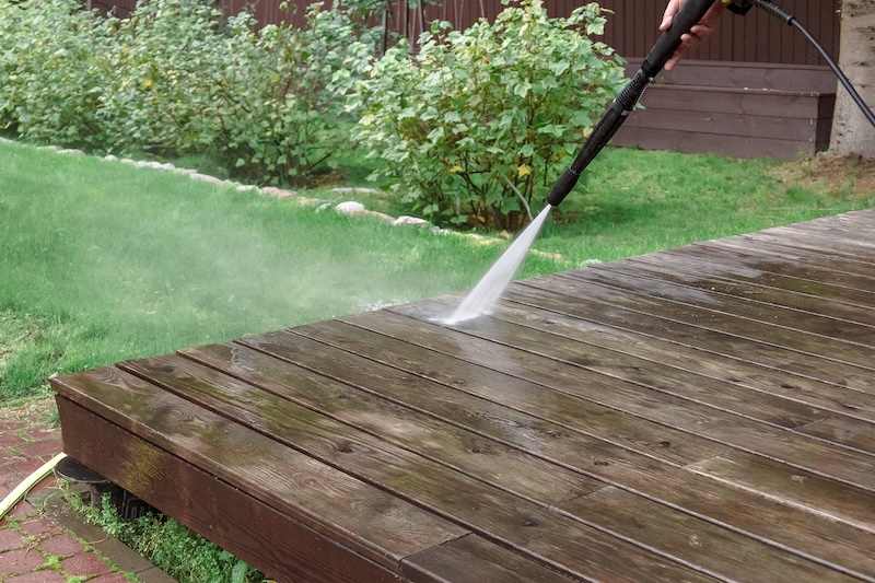 A Person Uses A Pressure Washer To Clean A Wooden Deck. The Stream Of Water Contrasts With The Green Grass And Bushes In The Sunny Backyard.