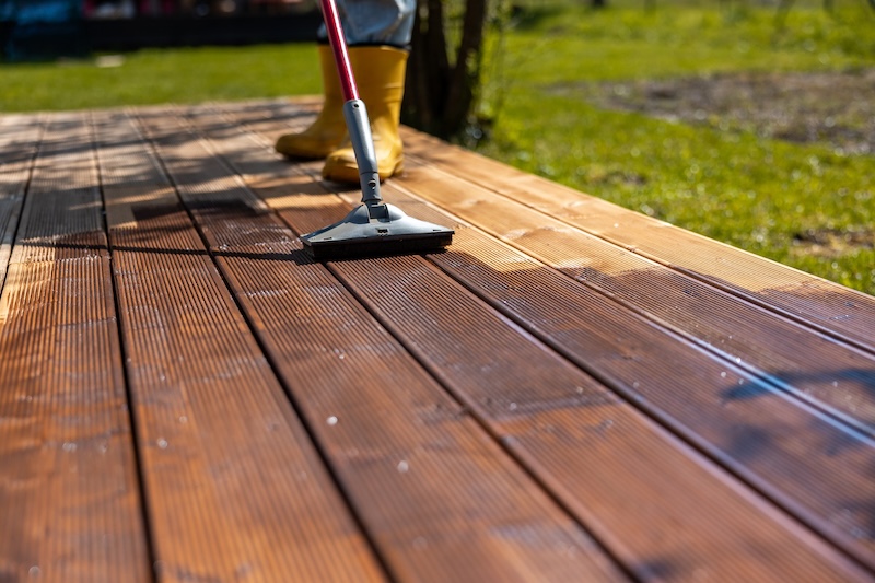CloseUp Of A Person Wearing Yellow Boots Using A Brush To Clean A Wooden Deck. The Deck Appears Wet, With Grass Visible In The Sunlit Background.