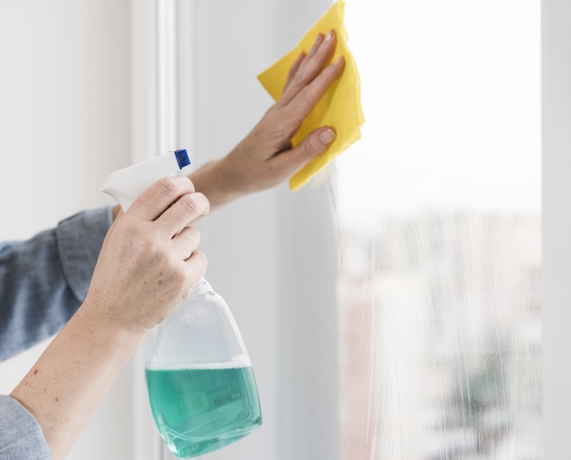 Hands Clean A Window With Blue Spray And A Yellow Cloth. The Background Is Blurred, Suggesting An Indoor Setting. The Scene Conveys Tidiness And Care.