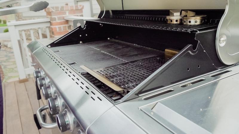 A Clean, Shiny Stainless Steel Grill With A Partially Open Lid And Visible Dials, Set On A Wooden Deck With A Blurred Brick Wall In The Background.