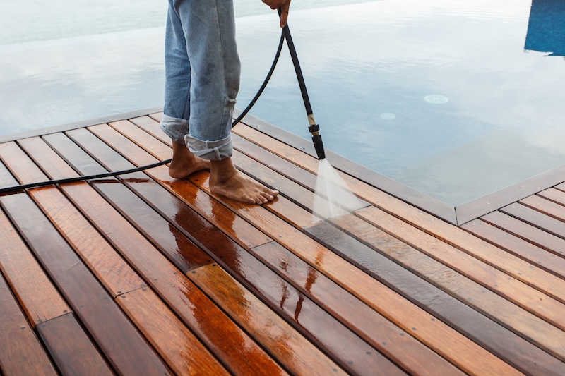 A Person In RolledUp Jeans Is Pressure Washing A Wooden Deck By A Calm Pool. The Scene Conveys A Sense Of Cleanliness And Tranquility.