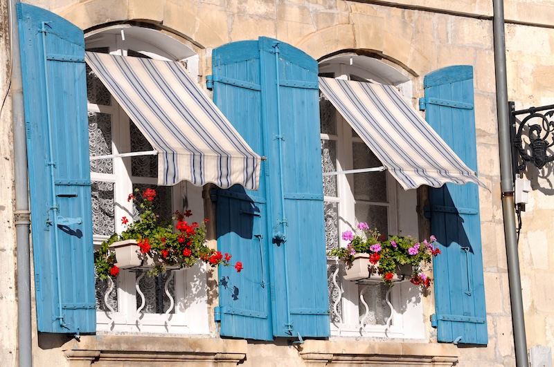 Drei Fenster Mit Leuchtend Blauen Fensterläden Und Gestreiften Markisen Auf Einem Steingebäude, Jedes Mit Blumenkästen Mit Roten Und Rosa Blüten. Helle, Fröhliche Atmosphäre.