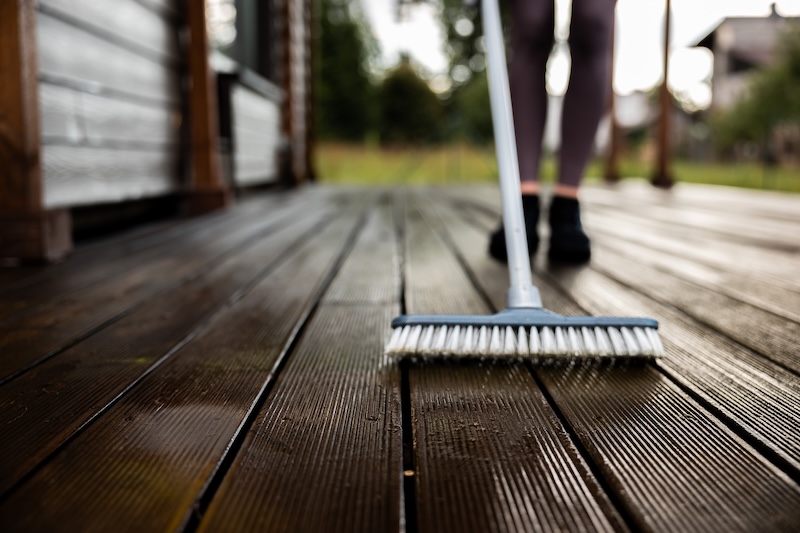 A Person Cleaning A Wet Wooden Deck With A Brush, Wearing Dark Leggings And Shoes. The Background Is Blurred, Giving A Focus On The Deck And Brush.