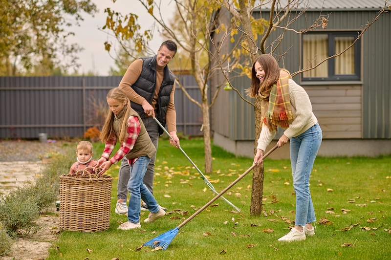 Working Mood Smiling Young Man Pretty Woman Warm Clothes Raking Leaves Lawn Girl With Little Brother Pouring Into Basket Garden Near House