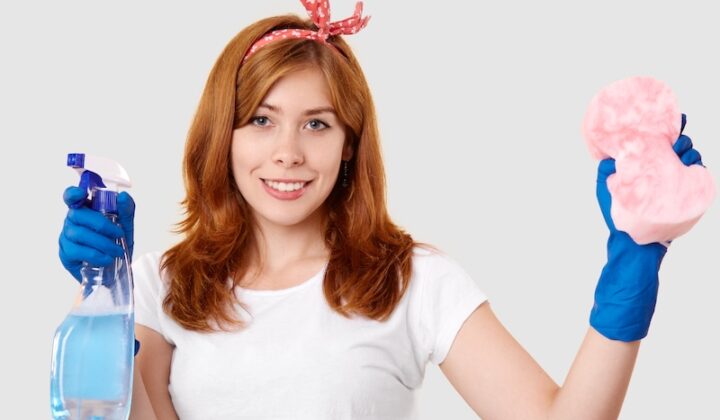 A woman holds a cleaning product in one hand and a sponge in the other, ready to clean a surface.