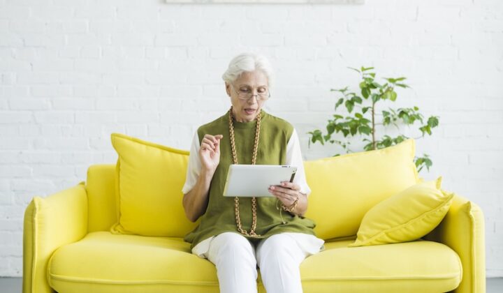 Eine ältere Frau sitzt auf einem gelben Sofa und hält ein Tablet in der Hand.