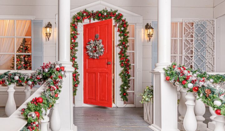 Eine rote Tür auf einer weißen Veranda, geschmückt mit einem Kranz und Girlanden.