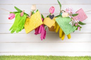 Colorful cleaning supplies hang on a clothesline with tulips and flowers, set against a white wooden background, evoking a cheerful, fresh spring vibe.
