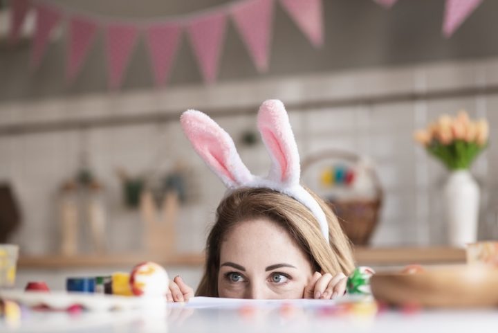 A woman wearing pink bunny ears peeks over a table, eyes wide with curiosity. Decorated eggs and pastel bunting suggest a playful Easter theme.