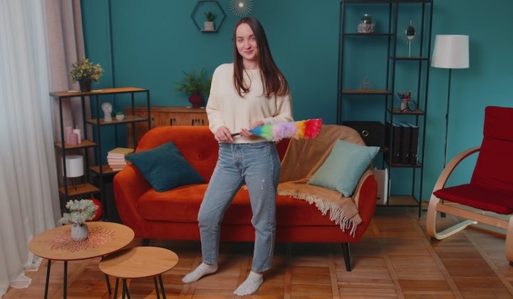 Happy one young woman dusting cleaning furniture caring for hygiene using colorful duster in living room. Girl housekeeper wiping dust from sofa and table at home. Housework and domestic life concept
