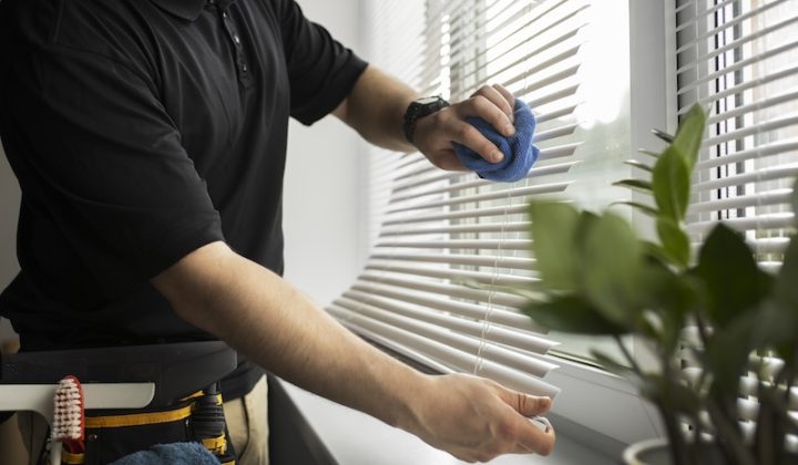 A person in a black shirt cleans a window with a blue cloth beside white blinds. A potted plant and cleaning supplies are visible, conveying tidiness.