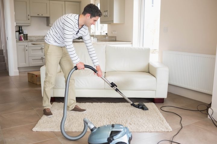Man in his twenties cleaning area rug with canister vacuum in living room, copy space. Modern, home, lifestyle, cleaning, interior, minimalism, productivity
