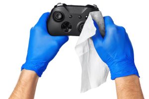 Hands in blue gloves clean a black game controller with a white cloth, set against a plain white background, conveying cleanliness and care.