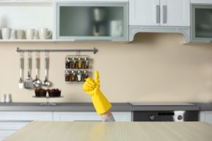 A yellow-gloved hand gives a thumbs-up, emerging from behind a kitchen counter. The modern kitchen has utensils and spice jars on the wall, creating a humorous tone.