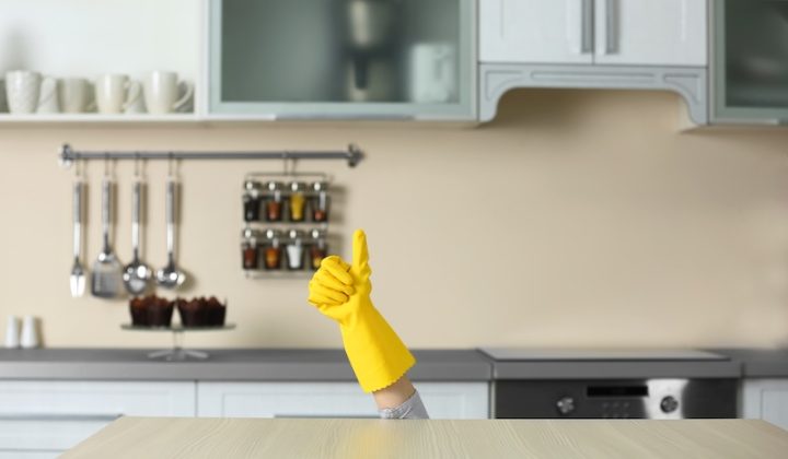 A yellow-gloved hand gives a thumbs-up, emerging from behind a kitchen counter. The modern kitchen has utensils and spice jars on the wall, creating a humorous tone.