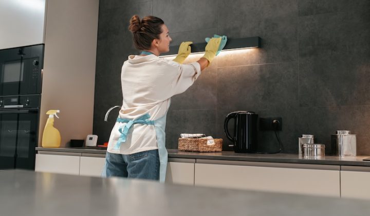A person wearing yellow gloves and an apron cleans a kitchen hood under soft lighting. A basket, kettle, and cleaning supplies are visible on the counter.