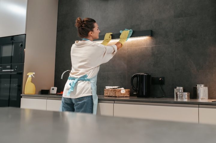 A person wearing yellow gloves and an apron cleans a kitchen hood under soft lighting. A basket, kettle, and cleaning supplies are visible on the counter.