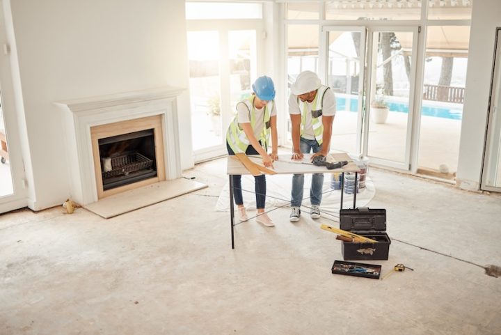 Two construction workers in hard hats and vests review blueprints on a table in a bright, unfinished room with a fireplace and large windows.