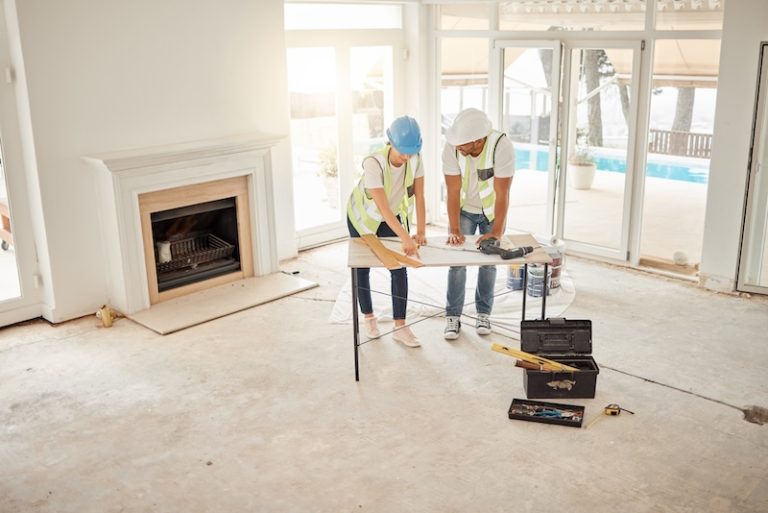Two construction workers in hard hats and vests review blueprints on a table in a bright, unfinished room with a fireplace and large windows.