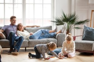 A relaxed family scene in a bright living room. A couple lounges on a gray sofa, while two children draw on the floor, conveying warmth and harmony.