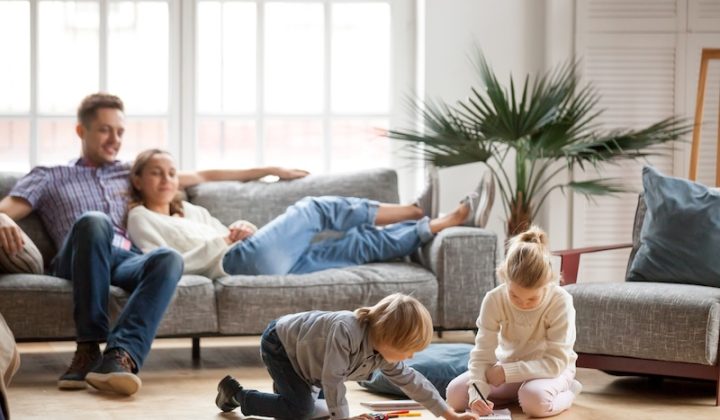 A relaxed family scene in a bright living room. A couple lounges on a gray sofa, while two children draw on the floor, conveying warmth and harmony.