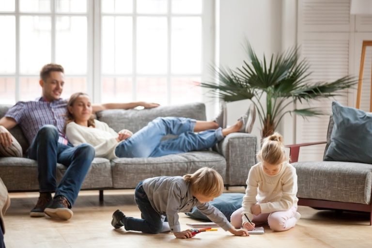 A relaxed family scene in a bright living room. A couple lounges on a gray sofa, while two children draw on the floor, conveying warmth and harmony.