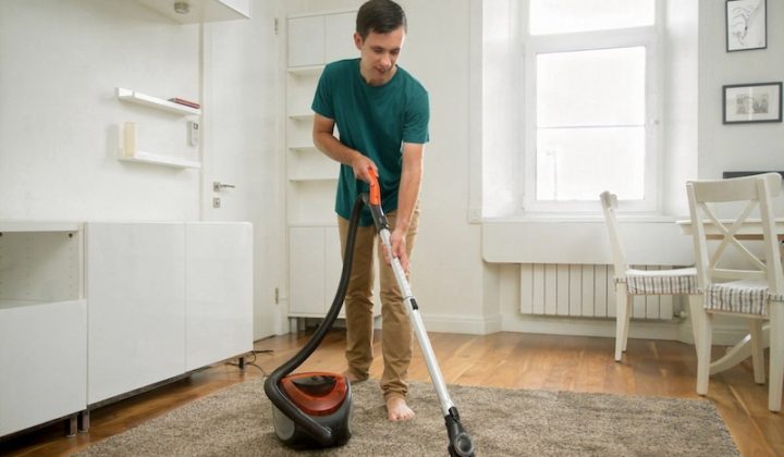 A Man In A Green Shirt Vacuums A Carpet In A Bright, Minimalist Room. The Space Has White Walls, Wooden Floors, And Simple Furniture, Conveying Cleanliness.