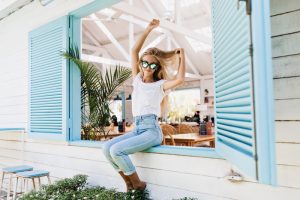A woman sits cheerfully on a window ledge, framed by light blue shutters. She's wearing sunglasses, a white shirt, and jeans, with hands in the air.