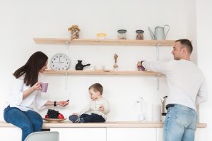 A family in a bright kitchen; a woman offers fruit to a child on the counter, while a man reaches for a mug. Shelves with jars and decor above them.