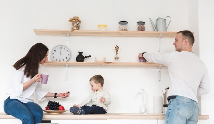 A family in a bright kitchen; a woman offers fruit to a child on the counter, while a man reaches for a mug. Shelves with jars and decor above them.
