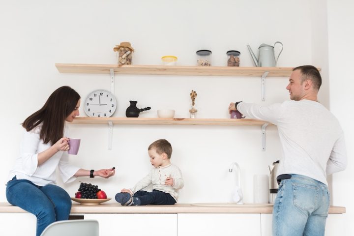 A family in a bright kitchen; a woman offers fruit to a child on the counter, while a man reaches for a mug. Shelves with jars and decor above them.