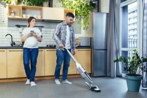 A man vacuums a modern kitchen with wooden cabinets. A woman stands nearby holding a smartphone. Green plants and natural light create a calm atmosphere.