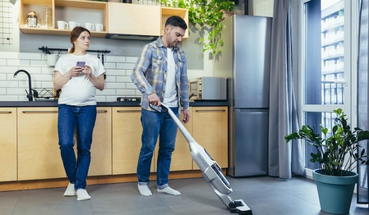 A man vacuums a modern kitchen with wooden cabinets. A woman stands nearby holding a smartphone. Green plants and natural light create a calm atmosphere.