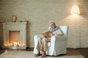Elderly woman in a cozy living room, reading a book on a white armchair. Lit candles fill the fireplace, creating a warm, peaceful atmosphere.