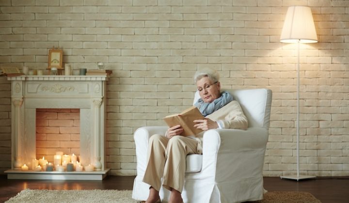 Elderly woman in a cozy living room, reading a book on a white armchair. Lit candles fill the fireplace, creating a warm, peaceful atmosphere.