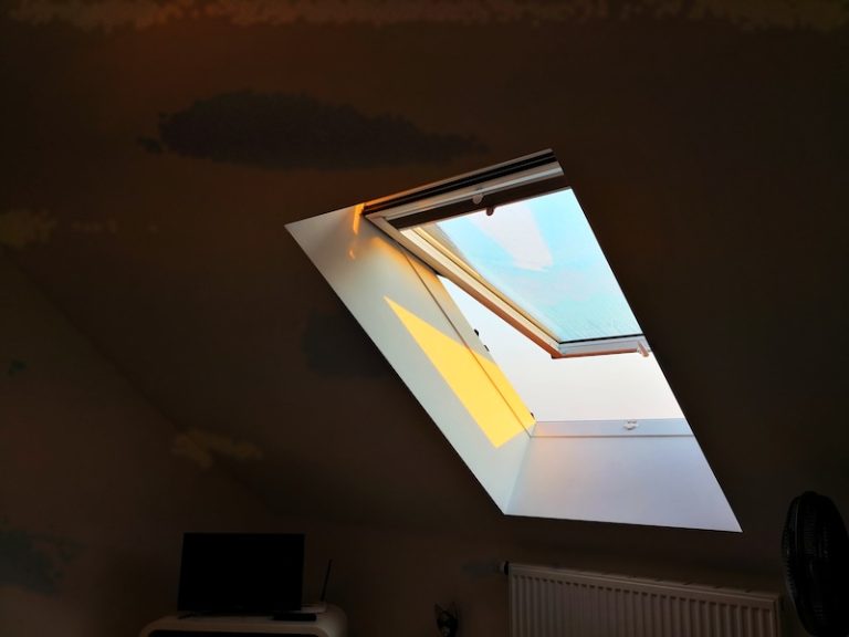 Sunlight streams through an open skylight, casting a warm glow on a dim attic room. A small TV and radiator are partially visible in the shadows.