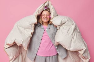 positive-cheerful-young-woman-wrapped-duvet-has-happy-expression-after-healthy-sleep-wakes-up-good-mood-wears-comfortable-pajama-makes-hairstyle-with-rollers-poses-against-pink-background